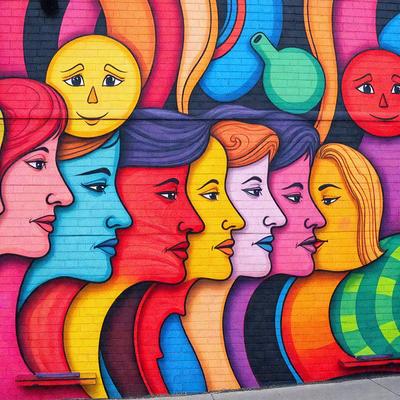 The primary subject of the image is a mural featuring a diverse group of people with different colored faces. The mural is painted on a brick wall, and the people are depicted in various positions and orientations. The setting is an urban environment, with the mural likely located in a public space. The visual style is a combination of digital art and illustration, creating a vibrant and engaging scene. The colors and mood of the mural are lively and energetic, with the different colored faces adding a sense